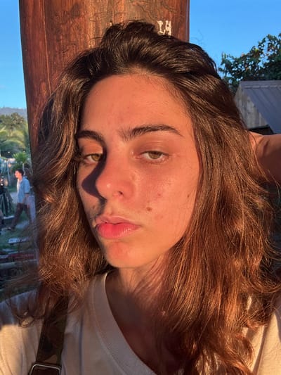 A young adult female is depicted centrally in an outdoor setting in Ban Mae Ping, Thailand. Her medium brown, wavy hair frames a medium skin tone with visible facial blemishes. Illuminated by strong, warm light from the right (viewer's left), pronounced shadows fall on her left side and beneath her nose. Her green-brown eyes are partially closed, and her lips are closed in a neutral expression. She wears a light-colored top, with a camouflage-patterned strap and metallic buckle visible on her right shoulder. The background to her left features a dark wooden post, beyond which a male figure in a light blue shirt and dark pants walks away. Further behind, green foliage, a partial structure, and a clear blue sky are visible. To her right, additional green foliage and a building roof are present against the clear blue sky.