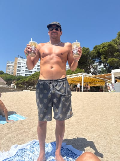 A man stands on a sandy beach in Platja d'Aro, Spain, holding two drinks. He is shirtless, wearing swim trunks, sunglasses, and a baseball cap. The beach has a bright, sunny atmosphere with a few buildings and trees in the background. The man is smiling and appears to be enjoying himself.