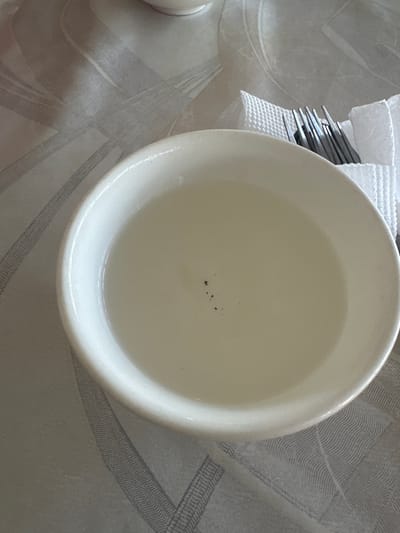 A white bowl of clear liquid sits on a textured tablecloth. Inside the bowl, a small cluster of dark specks is visible. To the right of the bowl, a fork rests on a white napkin. The environment appears to be indoors, possibly a dining setting. The location in the city of Kara-Suu, Kyrgyzstan, is not discernible from the image itself.