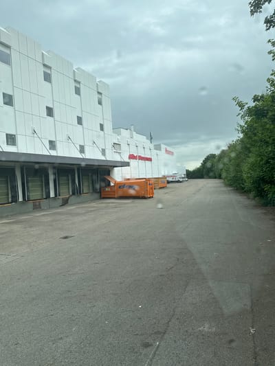 An exterior, eye-level, medium shot captures a loading dock area in front of a modern white building with a corrugated facade and numerous square windows. The building has a prominent red sign that reads "Möbel Wassermann." To the left, several large orange industrial dumpsters are parked near the loading dock doors. A white delivery truck is visible further down the paved area. The scene is flanked by a dense line of green trees on the right and a slightly overcast, cloudy sky above. The paved area appears to be an asphalt lot with some visible water marks and a storm drain. The image suggests a commercial or industrial setting, possibly a furniture warehouse or store, located in Memmingen, Germany.