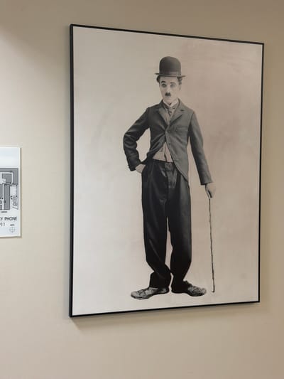 A framed, black and white photograph of actor Charlie Chaplin, dressed in his iconic "Tramp" persona, is displayed on a plain, light-colored wall. Chaplin is wearing a bowler hat, a suit jacket, a vest, a collared shirt with a tie, and loose-fitting trousers. He is holding a cane in his right hand and has his left hand on his hip. His expression is characteristic of his silent film performances, with exaggerated eyebrows and a small mustache. To the left of the photograph, a portion of a white document with a diagram and text is visible, appearing to be a floor plan or map. The lighting suggests an indoor environment, likely a public space or hallway. This photograph is located in the city of Burbank, United States of America.