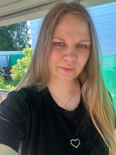 This is a selfie of a woman with long, blonde hair. She is wearing a black t-shirt with a small, gold heart pendant on the left side of her chest. Her eyes are blue-grey, and she has light skin with a few freckles visible on her nose and cheeks. She appears to be outdoors, with a green, translucent roof visible above and behind her. Trees and a white and green building are visible in the background. The lighting suggests it is daytime, likely sunny. The image seems to be taken in a casual, outdoor setting, possibly a porch or patio. The woman's expression is neutral. The text "Лотошино, Russia" is provided as location context.