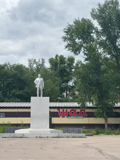 A silver statue of a man in a suit stands on a pedestal in front of a building with a striped facade. The building has a large red logo with white lettering that appears to say "ЕДА," and smaller text below it reads "СОЦИАЛЬНЫЙ СУПЕРМАРКЕТ" (Social Supermarket). Trees with lush green leaves surround the statue and the building. A paved area is visible to the right of the scene. The sky is overcast with clouds. The statue appears to be a memorial of some kind, with a dark plaque visible behind it, and flowers at its base. The overall scene suggests an outdoor public space in a Russian town. The time of day appears to be daytime, given the lighting. The weather is cloudy.