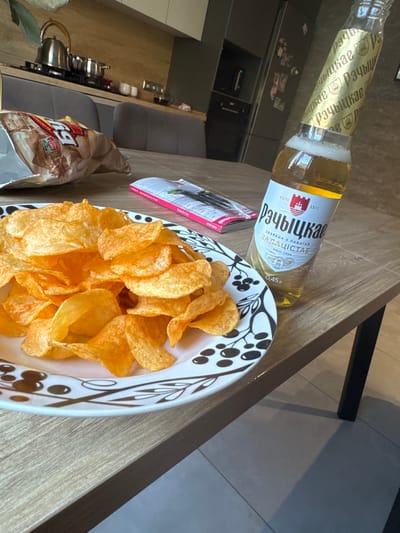 An indoor, eye-level, medium shot captures a portion of a light brown wooden table, bearing a plate full of golden-brown potato chips and a bottle of beer. The plate is white with a black and white abstract pattern around its rim. The potato chips are piled high on the plate, some slightly overlapping. Next to the plate, a clear glass bottle of beer stands at an angle. The beer is a pale yellow color with a frothy head visible at the neck of the bottle. The bottle has a gold label with text and a crest. In the background, a kitchen area is visible, with a silver kettle and pots on a stove, and a refrigerator. The scene appears to be set in a residential kitchen in Maladzyechna, Belarus.