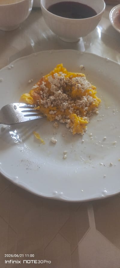 This is a top-down, medium shot photograph of a white plate with scrambled eggs and crumbled cheese on it. A fork is inserted into the eggs. The plate is on a reflective, light-colored surface, possibly a table. In the upper portion of the image, two white bowls are visible, one containing a dark liquid, likely a beverage. The lower portion of the image shows text overlaying the scene, indicating the date and time "2026/06/21 11:00" and the device "Infinix NOTE 30Pro". The scene is devoid of any discernible context related to the city of Sernovodskoye, Russia.