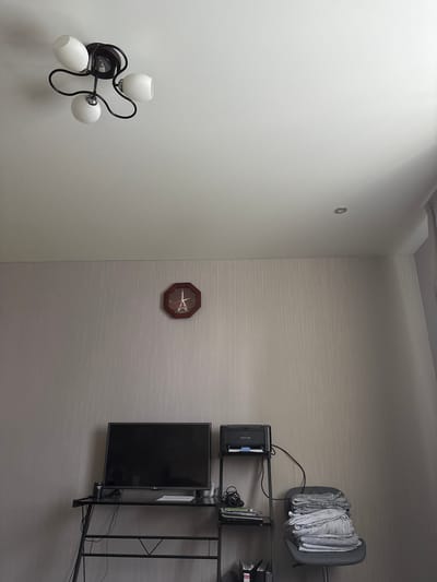 The image depicts an indoor scene, likely a domestic setting, possibly in the city of Kinel'-Cherkasy, Russia. In the upper portion of the frame, a ceiling light fixture with three white glass shades on black metal arms is visible. Below this, a wall features a decorative octagonal clock with a dark wooden frame, displaying Roman numerals and a central image of the Eiffel Tower. Further down, a black metal desk or shelf unit holds a black flat-screen television on the left. To the right of the television, a small black printer is positioned on a higher shelf. To the right of the printer, a gray office chair is stacked high with folded gray linens, possibly towels or bedding. The walls appear to be covered in textured wallpaper. The overall lighting suggests an interior environment with ambient light. There are no people or animals present in the scene.
