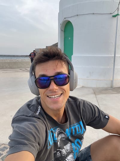 In a selfie taken outdoors in Cartagena, Spain, a man wearing grey over-ear headphones and reflective sunglasses smiles broadly. He is seated on a paved area, with a white cylindrical structure featuring a green arched doorway to his right. In the background, a body of water with a distant shoreline and a few figures can be observed. The man is wearing a dark grey t-shirt with blue text and an image on the front, and dark patterned shorts. The sky is overcast.