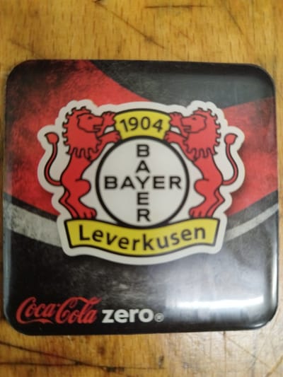 The image shows a square coaster with a dark background featuring a stylized red and black pattern. In the center of the coaster is a circular emblem with the Bayer logo. The emblem is outlined in white and contains the letters "B", "A", "Y", "E", "R" arranged vertically. Above the emblem, a yellow banner displays the number "1904". On either side of the emblem are stylized red lions. Below the emblem, a yellow banner reads "Leverkusen". In the bottom left corner of the coaster, the Coca-Cola Zero logo is visible in red. The coaster is placed on a wooden surface, which appears to be a table. The lighting is overhead, casting reflections on the coaster's surface. There are no people or actions depicted in the image. There is no information available to determine its location in Bremerhaven, Germany.