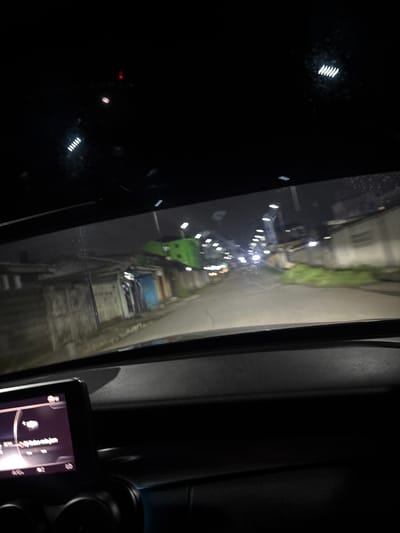 The image depicts a view from inside a vehicle, possibly a car, at night in Rumuomasi, Nigeria. Through the window, a narrow street is visible, illuminated by streetlights. A green dumpster is positioned on the left side of the road. Several parked vehicles are visible down the street. The interior of the car is partially visible, including a dashboard with a mounted electronic device. The environment is dark, with the city lights providing the primary source of illumination. No people are visible in the image.