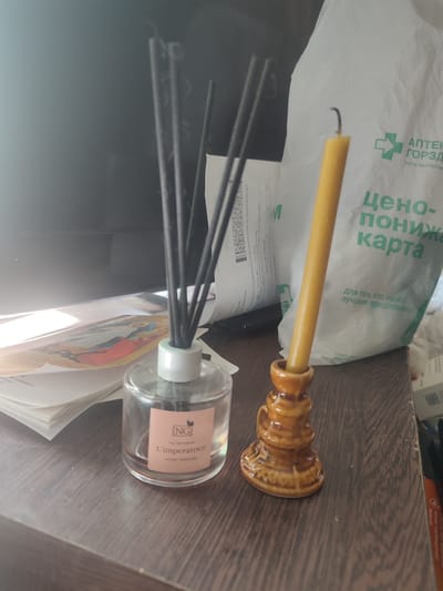 The image shows a close-up, eye-level view of a wooden surface with various objects on it. In the foreground, on the left, is a clear glass bottle containing multiple dark reeds, likely an aroma diffuser. A pink label on the bottle reads "NG L'imperatrice HOME PERFUME". To the right of the diffuser is a small, ornate, brown ceramic candlestick holding a single yellow candle. The candle appears partially burned, with its wick bent.
In the background, to the left of the diffuser, is a stack of papers, one of which has a colorful illustration visible. Behind the objects, a large white plastic bag with green text is partially visible. The text on the bag includes a green cross symbol and the words "АПТЕК" and "ЦЕНО- ПОНИЖАЮЩАЯ КАРТА", suggesting it may be from a pharmacy. The overall environment suggests an indoor setting, possibly a desk or table.
The location is not discernible from the visual information provided in the image.
