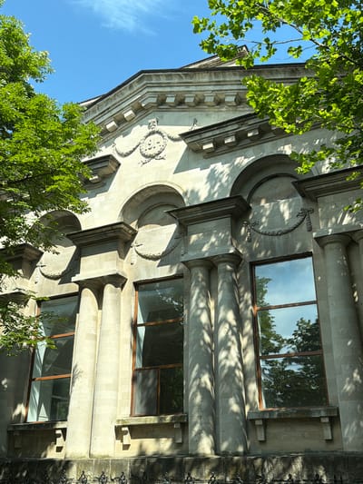 This is a close-up, angled shot of a light-colored stone building with intricate architectural details. The building features large, round columns and decorative elements, including a circular motif with what appears to be a stylized face or emblem above a recessed archway. There are several large windows with wooden frames, reflecting the surrounding greenery and sky.
Lush green trees with bright leaves are prominently visible in the foreground and partially obscure parts of the building, suggesting it is springtime or summer. The sunlight casts dappled shadows from the trees and architectural features onto the stone facade, indicating it is daytime, likely mid-morning or afternoon, given the angle of the light. The sky is a clear, bright blue with a few faint wisps of clouds.
The scene has a serene and somewhat grand feel, emphasizing the architectural beauty of the building and its integration with nature. There is no visible text in the image. The location is identified as Orhei, Moldova. No people or specific activities are depicted.