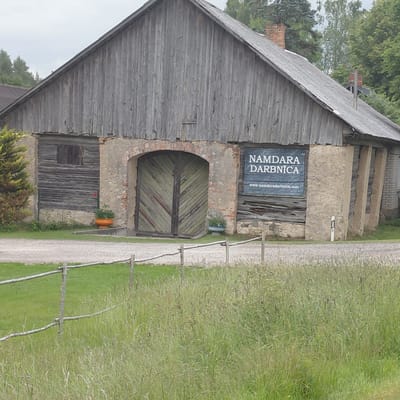 This is a vertical, slightly angled, medium shot of an old, rustic barn in Garkalne, Latvia. The barn appears to be constructed with weathered wooden planks on the left side, and stone and brick on the right. A large, arched doorway with weathered, green double doors is visible in the center of the stone section. Above the doorway, a sign is mounted on the wall. The text on the sign is in white capital letters: "NAMDARABNĪCA" followed by a website address "www.namdaradarnbnica.com". To the right of the main building, a gravel path runs alongside the barn, bordered by a simple wooden fence and lush green grass. The grass is tall and appears somewhat wild. A potted plant sits on a ledge next to the arched doorway. The roof of the barn is steeply pitched and covered in weathered grey wood. The overall impression is one of rural, historical charm, with the scene suggesting a quiet, possibly sunny day. The time of day is not definitively determinable, but the lighting suggests it could be late morning or early afternoon. There are no people visible in the image.