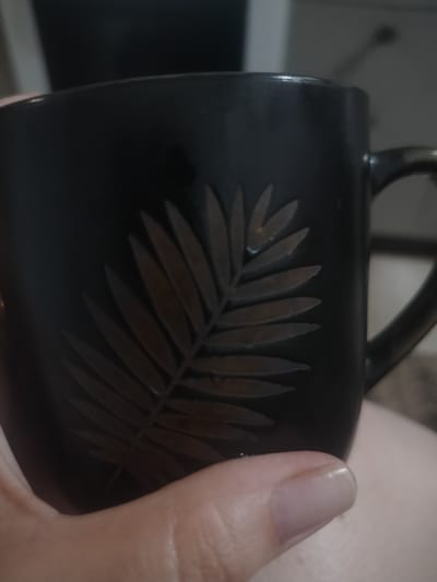 The image shows a close-up of a dark-colored mug with a raised leaf design on its surface. A hand is holding the mug, with a thumb and fingernail visible in the foreground. The mug is the primary object, and the hand is interacting with it. The background is dark and out of focus, providing no significant scene or setting details. There is no visible text in the image. The lighting appears to be indoor, as suggested by the soft illumination and lack of harsh shadows. The image does not convey any specific activity or event beyond the act of holding a mug. There are no indicators of time of day, weather, or specific location cues, beyond the provided "Yarovoye, Russia" context which is not visually evident. The overall mood is neutral.