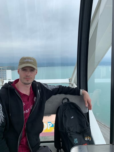 A man is pictured from the chest up, wearing a khaki-colored baseball cap with the word "Andyyou" on it, a red shirt, and a black zip-up jacket with white detailing on the sleeves. He is seated next to a black Puma backpack. The photo appears to have been taken inside an enclosed structure with large glass windows, suggesting a Ferris wheel or a similar observation attraction. Through the windows, a cityscape, a body of water, and an overcast sky are visible.
Based on the location, the cityscape and water may align with the city of Bichvinta (Pitsunda) in Abkhazia, but this cannot be definitively stated without more information.