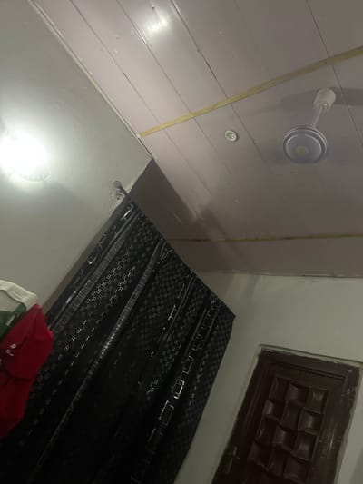 The image captures an indoor scene, likely a room, viewed from a low angle looking upwards. The ceiling is constructed of wooden planks, painted a light pinkish hue, with a visible yellow strip running horizontally across it. A white ceiling fan is mounted centrally on the ceiling. To the left, a white wall meets the ceiling, with a bright light source, possibly a bulb, illuminating the area. A section of a dark curtain with a repeating pattern of interlocking 'G' symbols and geometric shapes hangs diagonally from the ceiling to the floor. To the far right, a portion of a dark brown wooden door with a patterned surface is visible, set within a white wall. A stack of folded clothing, including red and green items, is partially visible in the lower left corner. The scene does not provide any specific visual cues to indicate its location within Pantinapu, Nigeria.