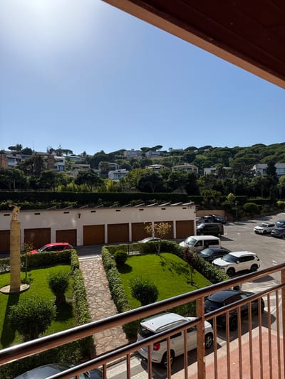This is a ground-level, eye-level shot of a residential area in Sant Feliu De Guíxols, Spain. The image is oriented vertically. A wooden railing in the foreground suggests the viewer is on a balcony or elevated walkway, looking down and across a steep hillside.
The hillside is densely covered with lush green trees, typical of the Mediterranean landscape. Scattered amongst the trees are several modern houses with white walls and flat roofs, indicating a contemporary architectural style.
Below the houses, a paved road runs parallel to the hillside, lined with parked cars. The cars are a mix of white and dark colors, with some appearing to be SUVs and sedans. To the right of the road, there are garages with wooden doors, suggesting a communal parking area or individual garage units. A neatly manicured lawn with bushes and trees is visible in front of the garages, with a stone pathway cutting through it. A small red car is parked to the left of the garages.
The sky is clear and a bright blue, suggesting a sunny day, likely in the late morning or early afternoon given the shadows cast by the sun. The overall impression is one of a well-maintained, upscale residential neighborhood in a hilly, coastal region. There is no visible text in the image.