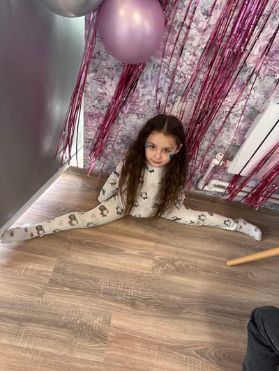 The image shows a young girl lying on the floor, with her legs spread in a split. She is wearing light-colored pajamas with a pattern of cartoon bears and hearts, and white socks with bear faces. The girl has long, dark, wavy hair and a blue mark on her cheek that looks like face paint. She is smiling at the camera, and her posture suggests she is showing off her flexibility.

The setting appears to be indoors, with a wooden floor. To the left of the girl, there is a wall with a floral wallpaper pattern, and pink metallic streamers hanging down, suggesting a festive occasion like a birthday party. A large pink balloon and a silver balloon are partially visible in the foreground, adding to the party atmosphere. Behind the girl, a white radiator is mounted on the wall. The lighting suggests it is daytime. The overall mood is joyful and playful. There is no visible text in the image. The location is Oseyevo, Russia.