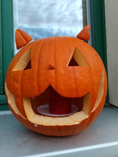 A single, orange, carved pumpkin is centered on a light-colored windowsill or ledge in Markkleeberg, Germany. The pumpkin features two triangular eye cutouts, a small inverted triangular nose cutout, and a wide, jagged mouth cutout exposing the hollow interior. Two distinct, pointed, ear-like shapes, also carved from pumpkin rind, are affixed to the top of the pumpkin. Inside the pumpkin, a red pillar candle rests on a white, circular dish. The background consists of a window with dark green framing, reflecting an overcast sky with white and light blue cloud formations. The surface beneath the pumpkin is a neutral, light gray color.