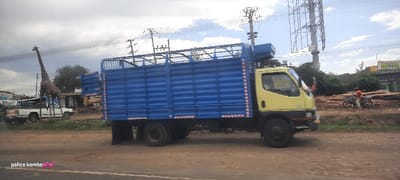 A blue and yellow truck is parked on a roadside with a tall, slender giraffe statue in the background near a white pickup truck. A motorcyclist rides near stacked logs further down the road. Power lines stretch across the sky and a large billboard stands in the distance. "police komba" text with pink hearts is visible at the bottom left.
