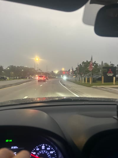 The image captures a street view from inside a stationary or slow-moving vehicle, with the dashboard and a partial speedometer indicating 0 MPH visible in the foreground. The scene is characterized by overcast and hazy weather conditions, resulting in low visibility and a uniformly gray sky. The road surface appears wet.

Multiple vehicles are present ahead on a multi-lane roadway; a light-colored SUV is directly in front of the viewer's vehicle. Streetlights are illuminated, casting a yellow glow, and vehicle headlights and taillights are active. Along the right side of the road, a series of American flags are displayed, followed by a large black vertical sign featuring the "Mitsubishi Motors" corporate logo. This identifies the establishment as a Mitsubishi Motors dealership. Parked vehicles are visible within the dealership lot behind the flags and sign. The left side of the road features trees and additional parked vehicles. Distant traffic signals show red lights. A human hand is partially visible in the lower-left section of the image. This specific scene is situated in the city of Doral, United States of America, consistent with the Mitsubishi Motors dealership located there.