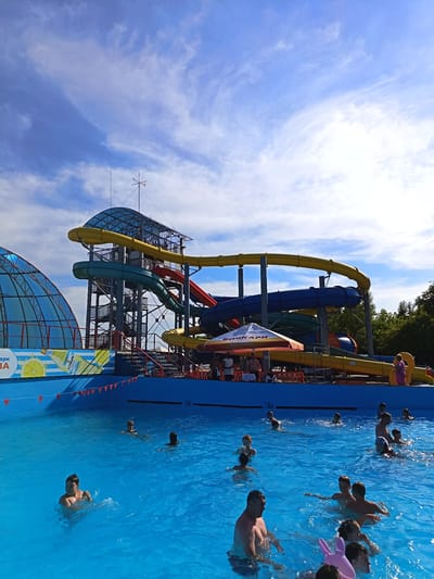 The image shows an outdoor water park in the city of Yarovoye, Russia. A large blue swimming pool fills the foreground, with numerous people of various ages swimming and playing in the water. In the background, a multi-story water slide structure with brightly colored tubes (yellow, blue, green, red) is prominent. A glass-domed structure with a metal frame is visible on the left. Several trees line the right edge of the frame. The sky above is a vibrant blue with scattered white clouds. A few people are visible standing on a deck area behind the pool, and a parasol is set up. Signs with text are present on the pool's retaining wall and on banners near the slides.