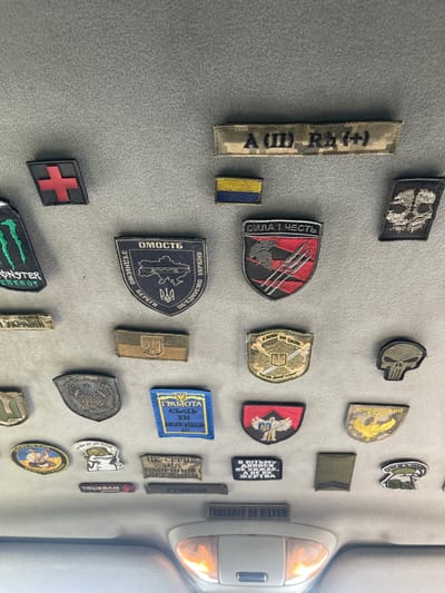 The image shows a ceiling panel of a vehicle, likely a car, covered in a variety of embroidered patches, commonly referred to as morale patches. The patches are attached to a gray, fabric-like surface. The collection of patches includes symbols, text, and imagery. Notable patches include a red cross on a black background, a skull design, a shield with Ukrainian national symbols, a patch with the Ukrainian flag, and several patches with Ukrainian text and military insignias. The lighting in the image suggests it was taken indoors, possibly during the day. There are no people visible in the image. The environment appears to be a vehicle interior, with a light fixture visible at the bottom of the frame. There is no information in the image to suggest the location is the city of Konopnytsia, Ukraine.
