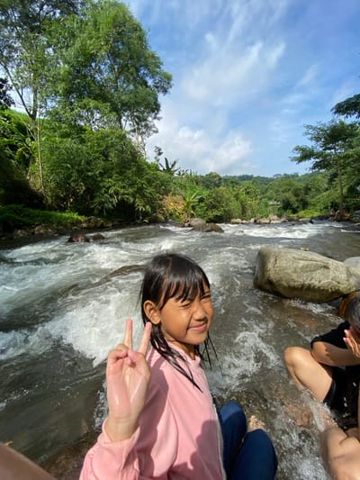 A young girl in a pink long-sleeved shirt lies partially submerged in a rushing river, her hair wet and plastered to her forehead. She smiles and makes a peace sign with her fingers. Other people are visible nearby, also enjoying the water. Lush green trees and vegetation line the riverbank on one side. Above, a blue sky with scattered clouds completes the outdoor scene.