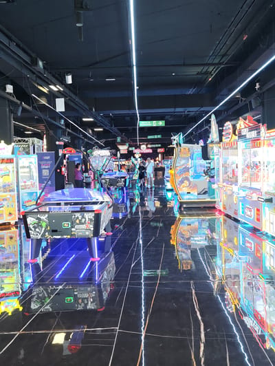 This image depicts the interior of a modern arcade and entertainment center in Nola, Italy. The space features a high ceiling with industrial lighting fixtures and ventilation systems. The floor is polished black marble with white veining, highly reflective and mirroring the bright neon lights and arcade machines.
The foreground is dominated by an air hockey table with blue LED lighting accents and colorful graphics. Behind it, a row of various arcade machines is visible, including claw machines with prizes inside and video game cabinets with vibrant digital displays. One machine prominently displays the text "GAME ORBIT" and instructions in Italian.
In the mid-ground, several people are present. A woman wearing a purple shirt is standing near a fire extinguisher, looking at one of the arcade machines. Further back, a man in a white t-shirt and shorts is walking with a child.
The environment is characterized by a playful and energetic atmosphere, suggested by the flashing lights, colorful machines, and the presence of patrons. The overall impression is a well-lit, clean, and technologically advanced entertainment venue.