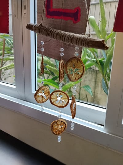 A close-up, eye-level shot captures a handmade wind chime hanging in front of a window. The wind chime is constructed from a rough, natural branch wrapped in twine. Dangling from the branch are several dried citrus slices, each separated by clear, faceted beads. One slice, at the bottom, appears to be a whole orange slice, while others are thinner, more irregular pieces. Behind the wind chime, a window reveals lush green foliage and a grey, weathered wall. The window frame is white, and to the left, a portion of a red rectangular object is visible, possibly part of another decoration. The lighting suggests it's daytime. The scene is indoors, with the window looking out onto an outdoor environment. It is not possible to determine the specific location within Ban Patan, Thailand.