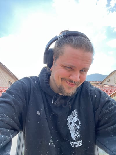 The image presents a head-and-shoulders portrait of an adult male, approximately 30-40 years old, situated outdoors under a bright, partly cloudy sky in Bogdašići, Montenegro. The subject faces the camera directly, displaying a slight smile. He has dark hair pulled back into a top-knot, a light-colored beard and mustache, and a silver-toned septum piercing in his nose. A subtle dark marking or tattoo is visible near his left eye. He is wearing black over-ear headphones and a black hooded sweatshirt, heavily splattered with white paint. The sweatshirt features a prominent white graphic print on the chest depicting a stylized, skull-like or gnarled face, with the text "DEVOTION TO LORD" legible beneath it. In the background, under the expansive sky, visible structures include a light-colored building with a terracotta-tiled roof to the viewer's left, and a second building with a similar roof and a mounted external utility unit to the viewer's right. Distant mountains or hills are discernible on the horizon.