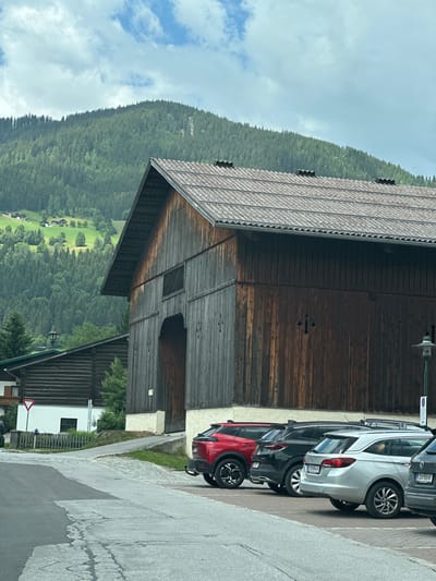 The image captures a cloudy day in Reitdorf, Austria, with a focus on a traditional wooden barn. The barn, constructed from dark wooden planks, features a large arched entrance. To the left of the barn, another building constructed from horizontal logs is visible, with a white section below it. A wooden fence runs along the front of the white section. In front of the barn, several cars are parked in a lot. These include a red SUV, a black SUV, a silver station wagon, and part of a dark colored vehicle. The cars are parked on a paved surface. In the background, a densely forested mountain slopes upwards, with scattered houses visible on its lower slopes. The sky is overcast with scattered clouds.