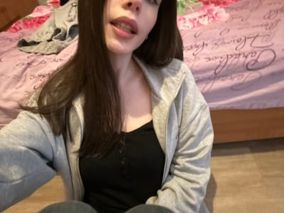The scene depicts a female individual in an indoor setting, likely a bedroom, in Volkhovets, Russia. The subject has long, dark hair and is looking towards the camera with her mouth slightly open, revealing some teeth. She is wearing a grey hooded sweatshirt with an open zipper over a black, ribbed top with three visible buttons. Her lower body, specifically the top of her legs, is visible, suggesting she is wearing dark-colored jeans. In the background, a bed is visible with a pink or light purple blanket/comforter featuring white floral and script patterns. A grey garment is loosely placed on the bed to the left of the subject. The floor visible at the bottom right consists of light-colored, wood-patterned planks. The lighting is even, suggesting an artificial light source or well-lit room.