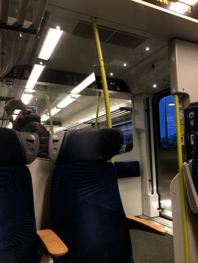 The interior of a train car is depicted, focusing on seating and overhead elements. Blue patterned seats and black headrests are prominent. A yellow handrail runs horizontally. Windows reveal a blue sky and trees outside. In the lower left, a passenger is visible, looking down at a device. Overhead lights illuminate the interior. The car appears clean and modern.
