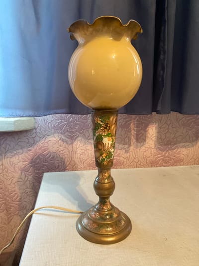 A vintage table lamp with a yellow, globe-shaped glass shade and a fluted rim sits on a white table. The lamp's base and stem are decorated with a painted nature scene featuring pine cones and green foliage. The lamp is plugged into an electrical outlet, and the cord runs across the table. In the background, a textured pink wallpaper is visible behind a blue curtain. The location is a domestic setting in Vasylky, Ukraine.