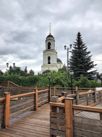 This media file captures an outdoor scene in Leshkovo, Russia, under an overcast sky. The foreground is dominated by a wooden walkway with railings, suggesting a path or a viewing platform. The walkway is constructed from planks and posts, with some sections appearing weathered. Beyond the walkway, lush green foliage forms a dense hedge, partially obscuring what lies behind.
Dominating the background is a white bell tower of a church, topped with a golden dome and a cross. The architecture suggests a religious building. Adjacent to the church is a tall, dark green conifer tree, which stands out against the cloudy sky. Several black lampposts with multiple lights are strategically placed along the walkway and within the greenery, indicating the area is likely used in the evening. The overall atmosphere is calm and somewhat somber due to the cloudy weather. No people are visible in the frame, and no text is present.