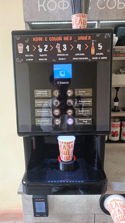 An outdoor coffee vending machine is situated in front of a stone wall that has text on it. The machine is black and has a touchscreen display. The display shows a cup and the word "Saeco". Below the screen, there are options for various drinks like hot chocolate, americano, cappuccino, and mocha. To the right, there are options for latte, americano, cappuccino, and banana raf. A paper cup with a red and white pattern is positioned in the dispensing area. A card reader is attached to the side of the machine. On the wall above the machine, there is text in Russian that appears to read "Coffee to go". The overall environment suggests a public area, possibly a street or a rest stop.