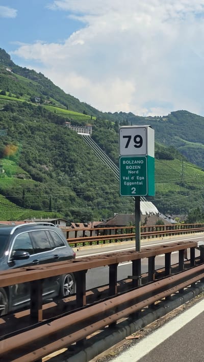 This image captures a highway scene in the vicinity of Bolzano, Italy. A dark-colored station wagon is moving on the highway, partially obscured by a brown metal guardrail in the foreground. To the right of the highway, a tall signpost displays road information. The top section of the sign has a white background with the number "79" in black. Below it, a green section reads "BOLZANO NORD" in white text, followed by "Val d' Ega Eggental" and the number "2" at the bottom. The background features a lush green mountainside dotted with buildings and terraced vineyards, with a partly cloudy sky overhead.