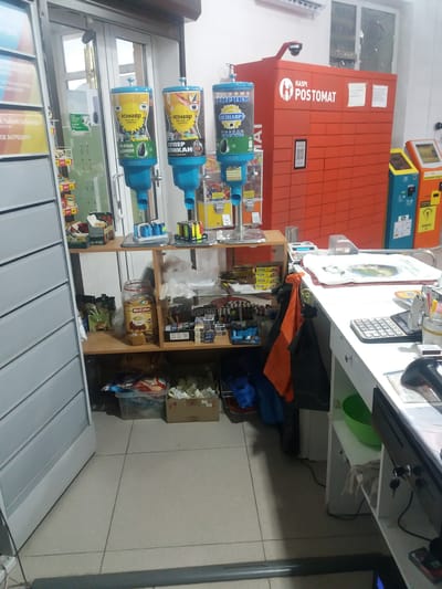 The image depicts the interior of a small convenience store or kiosk, likely located in the Бородулиха ауылдық округі, Kazakhstan, given the presence of Russian language signage and products. The scene is cluttered and typical of a retail environment.
In the foreground, to the left, a tall, grey shelving unit with multiple drawers is visible, its side facing the camera. In the center of the image, a white counter occupies the right half, on which several items are placed. These include a calculator, a scanner, a white cloth, a green bowl, and various papers. A black POS terminal with a touchscreen and a scanner is also prominent on the counter.
Behind the counter, on shelves, various products are displayed. These include packets of instant coffee, jars of snacks, and what appear to be lighters or similar small items. In the background, a large, bright red "Kaspi Postomat" parcel locker unit is a significant feature. To its right are two other electronic payment or vending machines, one yellow and one blue.
Further back, towards the left of the image, there are several dispensers for bulk goods, likely snacks or seeds, with blue bases and transparent containers holding colorful products. A security camera is mounted on top of the Postomat unit. The floor is tiled with light-colored square tiles, and the lighting suggests an indoor environment, possibly with fluorescent overhead lights. The overall impression is of a functional but somewhat unorganized retail space. There are no people visible in the image.