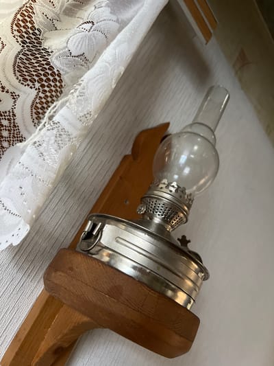A close-up, overhead shot shows a vintage oil lamp mounted on a wooden bracket against a textured white wall. To the left, a white lace curtain drapes the wall, creating a soft, patterned backdrop. The oil lamp, made of glass and silver-toned metal, features a clear glass reservoir and a decorative perforated metal collar. The wooden bracket is unfinished, with visible grain and a slightly rounded edge where the lamp rests. The lighting is soft, casting gentle shadows and highlighting the textures of the lace, wood, and metal. The scene evokes a sense of rustic charm or historical decor. No people are present. The location is the city of Nizhnee Sancheleevo, Russia.