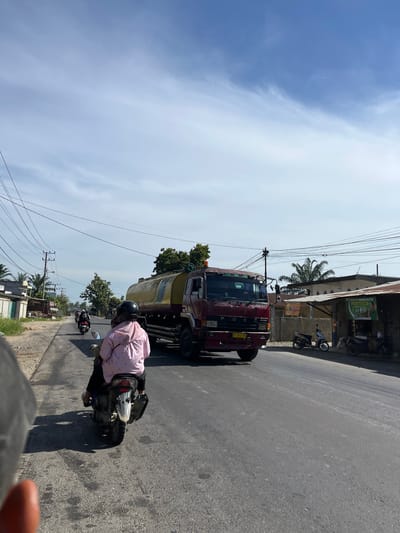 This media file depicts a street scene in Bukitrata, Indonesia, during the daytime under a bright, partly cloudy sky. The perspective is from someone riding a motorcycle, looking forward and slightly to the right.

The main subjects are a red truck with a large, pale yellow tank, and the person operating the motorcycle from which the photo is taken. The truck appears to be a water tanker. Another motorcycle is visible further down the road, also with a rider. The scene includes a paved road, a sidewalk with some vegetation, and buildings lining the street. Overhead, a complex network of electrical wires is prominent. One building has signage, one of which appears to be a banner with text and images.

The activity taking place is traffic on a street. The motorcycle the photographer is on is moving along the road, alongside the truck. The overall atmosphere is that of a typical day in a populated area with active road traffic. The bright sky suggests it is a sunny day. The presence of palm trees and the style of buildings and vehicles hint at a Southeast Asian location, specifically Indonesia as indicated by the location context.

Notable details include the license plate on the truck, which is partially visible and appears to have Indonesian characters and numbers, likely including "BL 8073 BD". The rider on the photographer's motorcycle is wearing a pink jacket and a black helmet. The truck has a yellow license plate with visible black text. The buildings have metal roofs and concrete structures. The sunlight casts distinct shadows on the road, indicating the time of day is likely late morning or early afternoon.