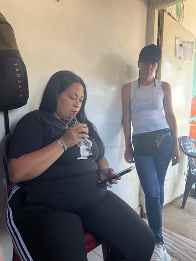 The image captures two adult females within a casual indoor or semi-outdoor setting in Quilmes Oeste, Argentina. On the left, a woman with long, dark hair is seated on a red chair or stool. She wears a black short-sleeved t-shirt with a light-colored graphic design and black ribbed pants with white stripes. Her right hand holds a mate gourd with a bombilla to her mouth, while her left hand holds a dark-colored smartphone, upon which her gaze is directed. She wears a silver-colored necklace with a pendant and a bracelet on her right wrist. To the left of her, a dark backpack with "RIP CURL" branding is suspended against a light-colored wall. On the right, a second woman stands, wearing a black baseball cap, a white sleeveless tank top, blue skinny jeans, and white athletic shoes. She has eyeglasses hanging by a cord around her neck and a black fanny pack positioned on her hips. Her posture suggests she is leaning slightly against the plain, light-colored wall. The background features primarily light-colored walls. To the right of the standing woman, an open doorway reveals a less defined interior space with some exposed framing and a hanging clear plastic object. A portion of a colorful mural or drawing is visible on the wall to the far right. A dark blue plastic chair with a patterned backrest is partially visible on the far right.