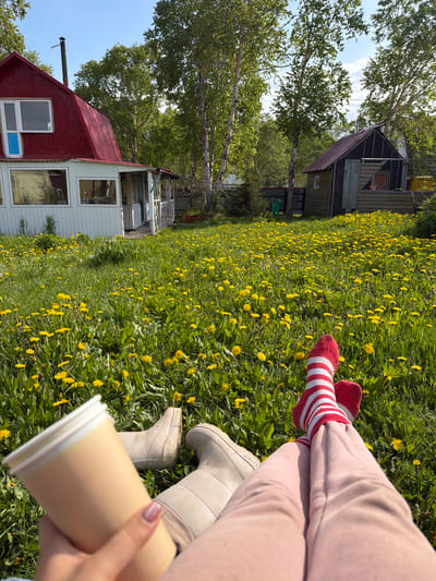In a grassy yard filled with yellow dandelions, two pairs of legs are visible, one person wearing red and white striped socks and pink sweatpants, the other wearing light-colored boots and partially obscured by a beige disposable cup. In the background, two houses stand under a clear blue sky. The house on the left has a red, sloping roof and white siding, with large windows. The house on the right is smaller, with a darker, sloping roof and wooden siding. Trees with green foliage surround the houses. The scene appears to be in Nagornyi, Russia, based on the context provided.