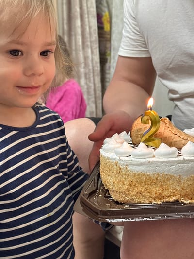 A young child with fair hair and a stripey blue and white shirt is positioned in the foreground. Their face is turned towards the viewer, and there is a small amount of white substance around their mouth. A cake with white frosting and a lit "2" candle sits above and behind the child. The cake is being held by a person with a white shirt. The background features a patterned fabric and a pink object, but these are not sharply defined. The location is Uva, Russia.