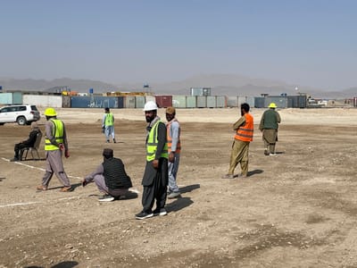 The image depicts an open, unpaved, dusty ground area in Pir Dust, Afghanistan, under a clear, pale blue sky with barren mountains in the distant background. A line of various colored shipping containers (blue, tan, yellow, green) stretches across the mid-ground, indicating a logistics or industrial site.
Approximately nine individuals are present, primarily adult males. Several wear personal protective equipment, including reflective safety vests (light green and orange) and hard hats (white and yellow) over traditional Afghan clothing (shalwar kameez).
* In the foreground right, a man with a beard wears a white hard hat and a light green vest, standing with a scarf around his neck.
* Adjacent to him, a man in an orange vest stands with arms crossed.
* Further right, a man in a yellow hard hat and dark green attire walks away from the camera.
* In the center foreground, a man in a dark plaid vest crouches, observing white lines painted on the ground.
* To the left, a man in a yellow hard hat and a light green vest walks left.
* A person in dark clothing sits on a black folding chair on the far left, with a white SUV (Toyota Land Cruiser type) parked behind them.
* Another man in a light blue outfit and light green vest walks towards the left background.
The white lines on the ground delineate a section of the dusty field. The scene suggests a gathering of personnel within a work or community context at an outdoor depot or construction-related site.