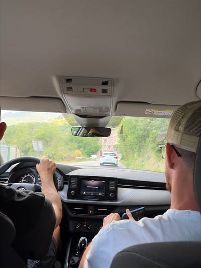 This is a first-person perspective image taken from inside a car traveling on a road in Svinjišta, Montenegro. The driver, wearing a black t-shirt, has their hands on the steering wheel. A passenger, wearing a white t-shirt and a baseball cap, is seated on the right, holding a blue smartphone. The car's dashboard is visible, including the infotainment system which displays a black screen with some Russian text, and the gear selector. Outside the car, a winding road is visible, flanked by lush green trees and hills. Another white car is seen ahead on the road. The sun is shining, casting shadows and illuminating the landscape.