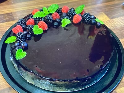 A circular, dark chocolate glazed cake, decorated with fresh raspberries, blackberries, blueberries, and mint leaves, sits on a black cake stand. The cake stand is placed on a wooden surface. The image is taken from a high-angle perspective, with the cake occupying the majority of the frame. There are no people or animals present. The scene appears to be indoors, likely in a kitchen or dining area. No specific location in Purkersdorf, Austria, can be identified from the image.