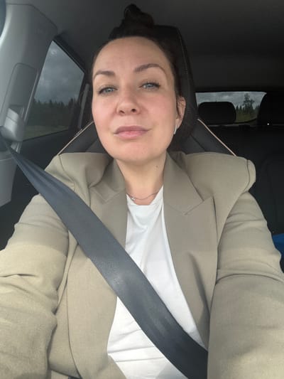 A woman is captured in a selfie taken from inside a car, likely in the passenger seat, in Kukushtan, Russia. She has dark hair, blue eyes, and a light complexion, exhibiting a calm and slightly smiling expression. She is wearing a white t-shirt, a light tan or beige blazer, and a delicate silver necklace with a small pendant. A black seatbelt is fastened across her chest.
Through the car window, an overcast sky is visible above a stretch of dark green trees, consistent with a rural or wooded setting. The interior of the car is dark, with visible seat upholstery and a headrest. Partially visible and inverted text that appears to read "AIRBAG" can be seen on the car's interior, possibly on a headrest or pillar. The time of day is daytime, characterized by cloudy or overcast weather.