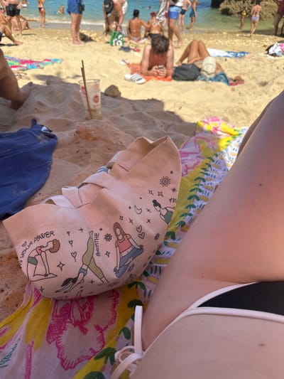 The image captures a sunny beach scene in Lagos, Portugal, from a low, personal perspective. In the immediate foreground, the right leg and hip of a light-skinned individual are visible, clad in a black bikini bottom with white tie details. Adjacent to this, a light beige fabric tote bag rests on a colorful beach towel featuring yellow, pink, and green patterns. The tote bag is illustrated with various women in yoga poses, alongside text including "OWN YOUR POWER" and "BREATHE." To the left, a dark blue garment is crumpled on the sand. A clear plastic cup with a red logo and two brown straws is partially buried in the sand behind these items.

The midground reveals a sandy beach populated with multiple individuals. One person lies prone on an orange towel, engrossed in a smartphone, with white Adidas slides near their head. Another individual is partially visible, lying supine with a hat over their face. Other beachgoers are scattered across the sand, either relaxing or moving towards the water.

The background features the turquoise-blue ocean, where several people are standing or wading. A distinct rocky outcrop is visible in the water on the far right. Sunlight illuminates the entire scene, casting shadows on the sand.