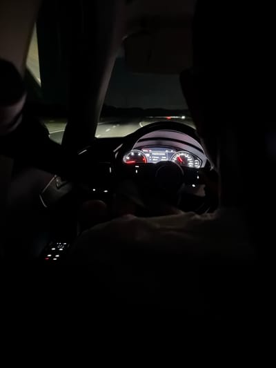 The image shows the interior of a car at night, as evidenced by the dark environment and illuminated dashboard. The dashboard displays a speedometer with a needle pointing to approximately 120 km/h, indicating the car is in motion. There are also other illuminated displays and controls visible. Two individuals are present, one in the driver's seat and another in the passenger seat, though their features are obscured by the darkness and framing. The road is visible through the windshield, with faint lights and a dark landscape in the distance, suggesting an outdoor setting, possibly a highway. The image is a low-light, personal photograph captured from inside the vehicle. There is no information within the image that indicates the specific location within the city of Aheloy, Bulgaria.