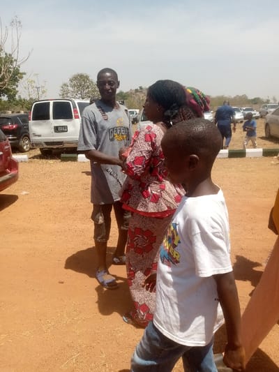 The image captures an outdoor scene in Kagoma, Nigeria, under bright daylight. The ground is unpaved, reddish-brown earth with sparse dry vegetation.
In the mid-ground, three individuals are prominent. On the left, an adult male stands facing right, wearing a grey short-sleeved t-shirt with a "DEFEND THE LAND" graphic and visible distressed areas or tears, dark capri-style shorts, and light-colored sandals. In the center, an adult female stands facing left, wearing a red, black, and cream patterned traditional dress and a dark head covering. The man and woman are positioned as if in conversation. In the right foreground, a young child stands with their back partially to the camera, wearing a white short-sleeved t-shirt with a colorful graphic on the front and blue jeans.
In the background, a white cargo van with tinted windows is parked directly behind the adult figures. To its left, a dark SUV is parked. Numerous other vehicles are visible parked further in the distance, suggesting a gathering or parking area. Low white and green painted concrete blocks or curbs are visible in the right mid-ground. The far background features low, vegetated hills under a hazy, light blue sky. Distant figures are visible, including an adult in dark attire and a child in blue and light pants. Shadows are visible on the ground, indicating a sunny environment.